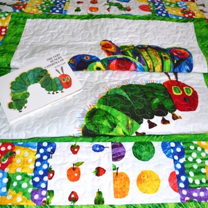 May include: A white quilt with a green border. The quilt features a patchwork design with colorful squares and circles. The center of the quilt features a large green caterpillar with red eyes and a book titled "The Very Hungry Caterpillar" by Eric Carle.