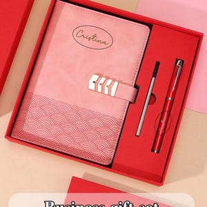 Puede incluir: Un juego de regalo de negocios rosa que incluye un bloc de notas con "Cristina" impreso, un bolígrafo y una recarga, todo presentado en una caja de regalo roja. El bloc de notas presenta un diseño de patrón de olas. El conjunto está etiquetado como "Customized content".