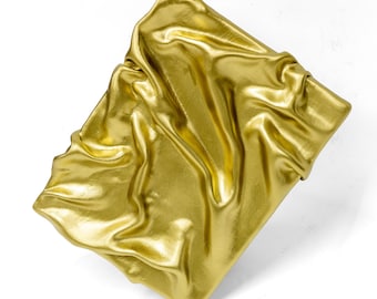 May include: A gold-coloured, rectangular object with a draped fabric appearance. The fabric has a shiny, metallic finish and is arranged in soft folds and wrinkles, creating a textured surface. The object is set against a white background.