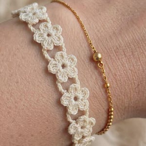 Könnte beinhalten: Ein cremefarbenes Häkelarmband mit einer Reihe verbundener Blumenmotive neben einem dünnen, goldfarbenen Metallkettenarmband mit einem kleinen kugelförmigen Perlendetail. Beide Armbänder werden zusammen getragen und zeigen kontrastierende Texturen.