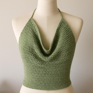 May include: A sage green crochet halter top with a cowl neckline, displayed on a mannequin. The halter straps are thin, and the body of the top has a textured stitch. The top is a solid colour. The garment is handmade.