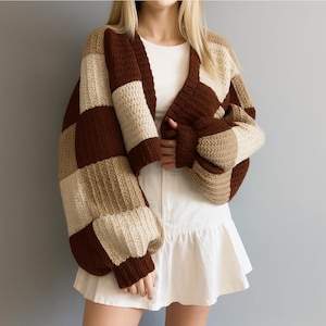 May include: A brown, beige, and cream-colored crochet cardigan with a checkerboard pattern. The open-front sweater has long, puffy sleeves and a V-neck. The garment is made of a chunky yarn and appears to be handmade.