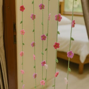 Op de afbeelding: Een decoratief gordijn gemaakt van gehaakte bloemen in roze en paarse tinten, geregen op groen garen. De bloemen zijn gelijkmatig verdeeld over de verticale strengen, waardoor een delicaat en grillig ontwerp ontstaat. Het gordijn hangt voor een neutraal gekleurde muur.