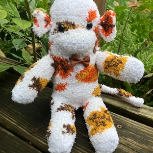 May include: A plush monkey toy crafted from white, orange, and brown mottled fabric. The stuffed animal features two round ears, black button eyes, and a small brown bow tie at the neck. It has long arms, legs, and a tail, with a soft, fuzzy texture.