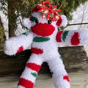 Könnte beinhalten: Eine handgefertigte Sockenaffenpuppe mit weihnachtlichem Thema. Die Puppe ist überwiegend weiß mit roten und grünen Akzenten, darunter eine rote Nase, Flecken und eine Schleife. Die Schleife hat ein festliches Muster.