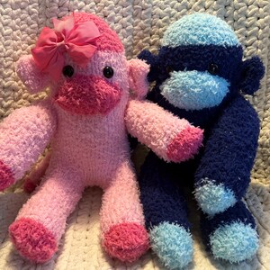 May include: Two handmade sock monkeys. One is pink with a pink bow, the other is navy blue and light blue. Both have black button eyes and are made of soft, fuzzy material. They are sitting on a cream-colored, textured surface.