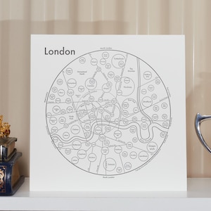 May include: A square white print featuring a circular black line map of London with various neighborhood circles and the text London in the top left corner. The print is displayed on a shelf between a stack of two books with a gold crown and a silver bowl.