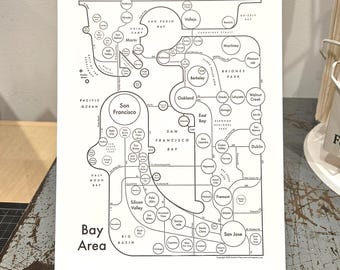 May include: A white rectangular print featuring a black line-art schematic map of the San Francisco Bay Area. The design uses circles to represent cities and locations, connected by thin lines, with the text Bay Area printed in the bottom left.