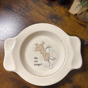 May include: Off-white ceramic plate with handles and a gold rim. The plate features a cartoon angel illustration with the text "Be an angel". The angel is blowing a trumpet. The plate is on a wooden surface.
