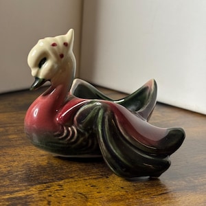 May include: A ceramic duck ornament with a cream head and a red and green body. The duck has a long neck and a curved beak. The ornament is likely a decorative item.