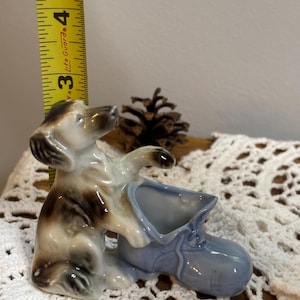 May include: A ceramic figurine of a dog with brown and white markings, interacting with a light blue shoe-shaped planter. The figurine is approximately 3 inches tall.