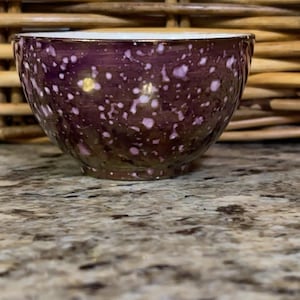 May include: A small, round ceramic bowl featuring a deep purple exterior with a speckled pattern of lighter pink and white spots. The interior of the bowl is smooth and white, contrasting with the dark, textured appearance of the outer surface.