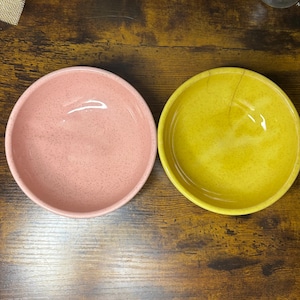 Könnte beinhalten: Zwei Keramikschalen, eine rosa und eine gelbe, stehen nebeneinander auf einer Holzoberfläche. Die Schalen haben eine gesprenkelte Textur und ein glänzendes Finish. Die rosa Schale befindet sich links, die gelbe rechts.
