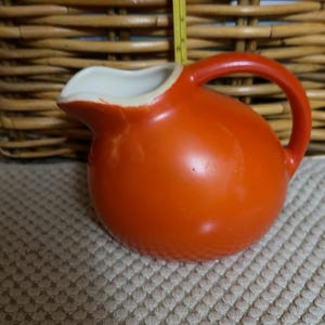 May include: A round, bright orange ceramic pitcher with a curved handle and a white interior. A yellow measuring tape is positioned vertically behind the pitcher, showing a height of approximately 4 inches from the base to the top rim.