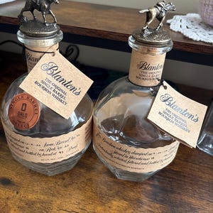 以下が含まれることがあります： Blanton's Single Barrel Bourbon Whiskeyの透明なガラス瓶が2本。各ボトルには、ブロンズ色の馬と騎手のフィギュアがトップに付いたコルク栓が付いています。ボトルにはテキストが記載された紙ラベルが付いています。