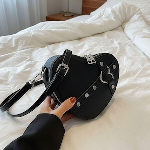 May include: Black heart-shaped handbag with silver hardware, including a zipper, buckles, and rivets. The bag has a top handle and a detachable shoulder strap. The bag is made of a smooth material.