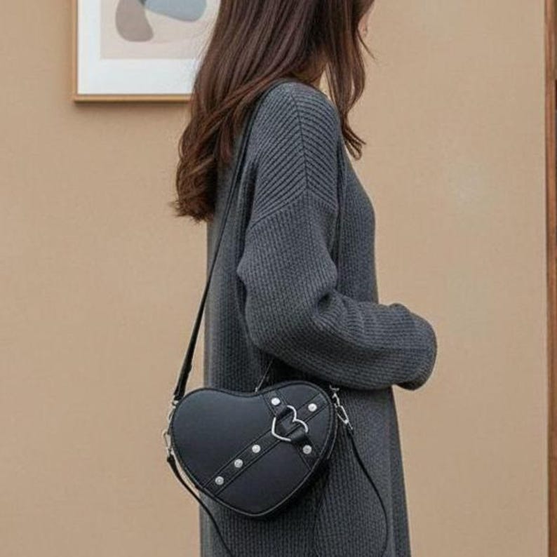 May include: Black heart-shaped crossbody bag with silver-tone hardware. The bag features a flap closure with a heart-shaped buckle and decorative studs. The bag is worn over the shoulder with a thin strap.