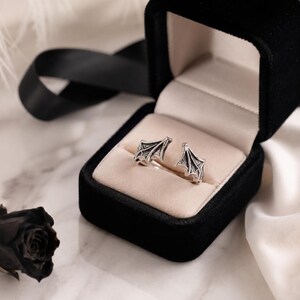 May include: A pair of silver-colored stud earrings shaped like bat wings, displayed in an open black velvet jewelry box with a cream-colored interior. The earrings have intricate detailing and are set against a backdrop of a black ribbon and a black rose.