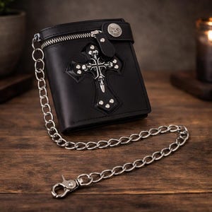 May include: Black leather wallet with silver cross and skull detailing. The wallet has a zip closure, chain, and skull button. The cross is decorated with silver studs. The wallet is on a wooden surface.