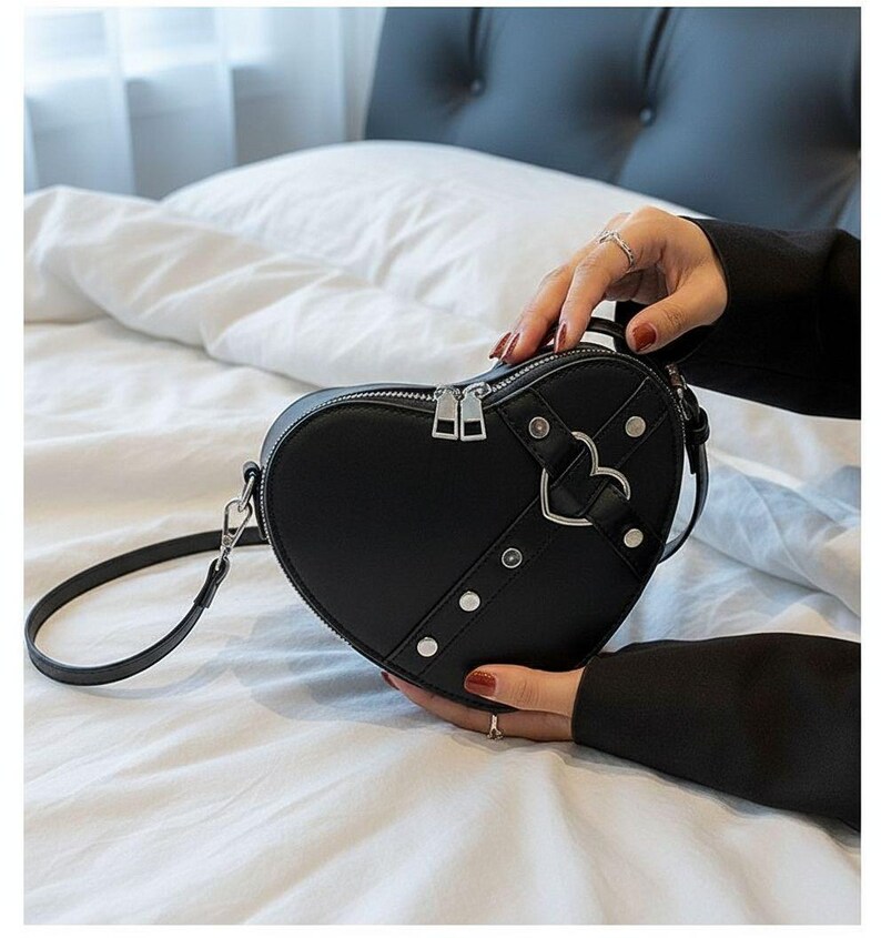 May include: Black heart-shaped crossbody bag with a silver zipper and hardware. The bag features a decorative heart-shaped buckle and silver rivets. The bag has a long, thin strap for shoulder or crossbody wear.