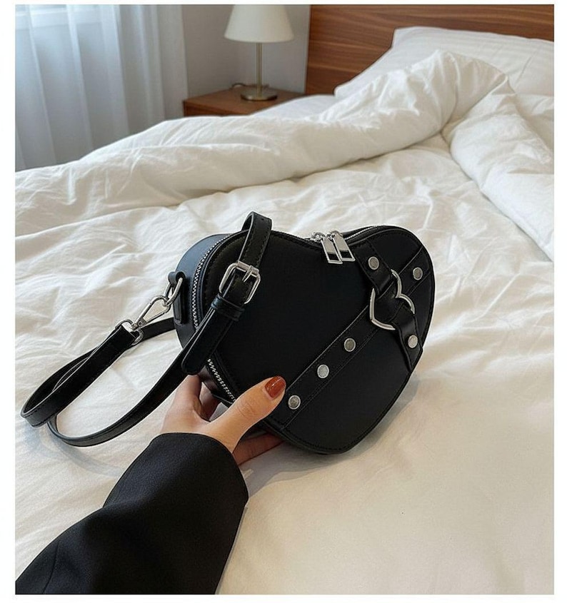 May include: Black heart-shaped handbag with silver hardware, including a zipper, buckles, and rivets. The bag has a top handle and a detachable shoulder strap. The bag is made of a smooth material.