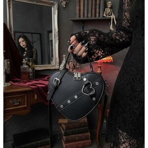 May include: Black heart-shaped handbag with silver hardware, including a zipper closure and a decorative heart-shaped accent. The bag has a shoulder strap and is studded with silver rivets. The bag is held by a person wearing a black lace sleeve.