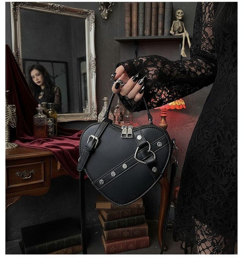 May include: Black heart-shaped handbag with silver hardware, including a zipper closure and a decorative heart-shaped accent. The bag has a shoulder strap and is studded with silver rivets. The bag is held by a person wearing a black lace sleeve.
