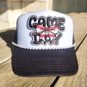 May include: A black and white trucker hat with the words "GAME DAY" in black lettering. A baseball-themed bow is centered on the front. The hat has a pearl bead trim along the brim.