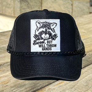 May include: Black trucker hat with a mesh back and a curved brim. The front features a white square patch with a raccoon illustration and the text "Sweet, BUT WILL THROW HANDS". A black chain detail accents the brim.