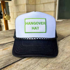 May include: A black and white trucker hat with the words "HANGOVER HAT" in green on a white patch. The hat has a black brim and a chain detail above the brim.
