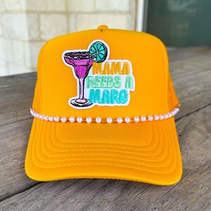 May include: A bright yellow trucker hat featuring an embroidered patch of a pink margarita glass with a lime slice. The text reads MAMA NEEDS A MARG in orange, green, and blue. A single strand of small white faux pearls lines the base of the crown.