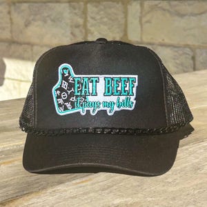May include: Black trucker hat with a mesh back and a curved brim. The front features a patch with the words "EAT BEEF it pays my bills" in teal and white, with a decorative border.