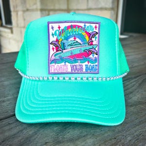 May include: A turquoise trucker hat with a white pearl beaded brim. The front features a patch with the words "Whatever Floats Your Boat" and a graphic of a boat with dolphins. The hat has a mesh back.