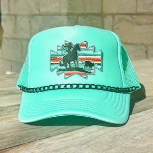May include: A turquoise trucker hat with a black chain detail and a mesh back. The front features a colorful embroidered patch with a cowboy on a horse and a cow. The hat is sitting on a wooden surface.