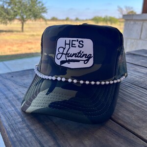 May include: A camouflage trucker hat with a white patch that reads "HE'S Hunting" in black script. The hat features a pearl-like beaded band across the brim. The hat is sitting on a wooden surface.