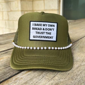 May include: Olive green trucker hat with a white patch that reads "I BAKE MY OWN BREAD & DON'T TRUST THE GOVERNMENT." The hat is decorated with a string of white pearls along the brim. The hat is sitting on a wooden surface.
