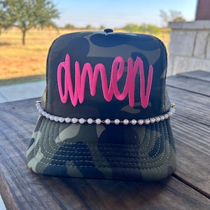 May include: A camouflage trucker hat with the word "amen" embroidered in bright pink script across the front. The hat features a pearl and gold chain detail along the brim. The hat is sitting on a wooden surface.