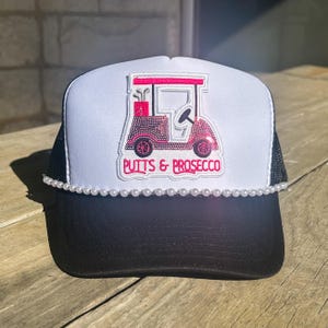 May include: A black and white trucker hat with a white front panel. The front features a pink golf cart design and the text "Putts & Prosecco". A string of white pearls adorns the brim.