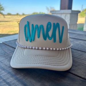 May include: Tan trucker hat with the word "amen" in teal script across the front. The hat features a pearl beaded band along the brim and a mesh back. The hat is sitting on a wooden surface.