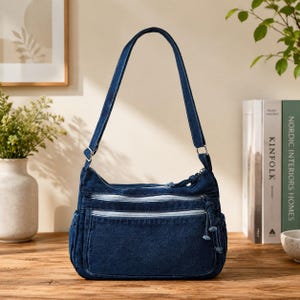 May include: A dark blue denim shoulder bag featuring a single adjustable strap, two horizontal zippered front pockets with silver-toned hardware, and side pockets. The bag has a structured rectangular base and a top zipper closure with knotted pulls.