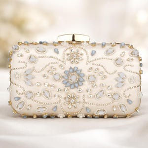 Puede incluir: Bolso de mano rectangular marfil con herrajes dorados. El bolso está adornado con cristales azul claro y transparentes, cuentas y diseños florales. El cierre es dorado. El diseño es elegante y adecuado para ocasiones especiales.