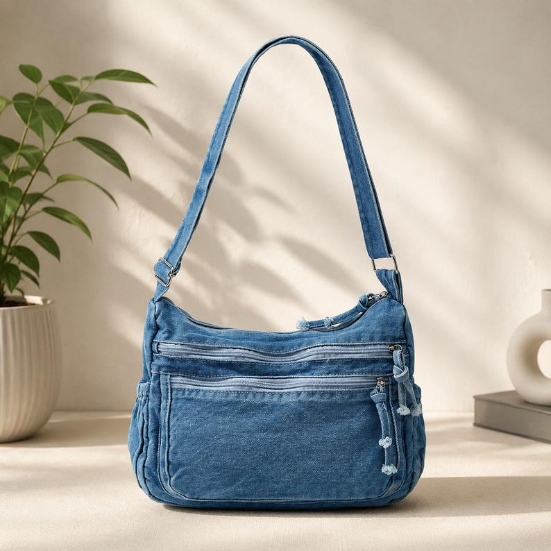 May include: A medium blue denim shoulder bag featuring a single adjustable strap, two horizontal zippered front pockets, and a main zippered compartment. The bag includes silver-toned metal hardware and knotted fabric zipper pulls on the front.