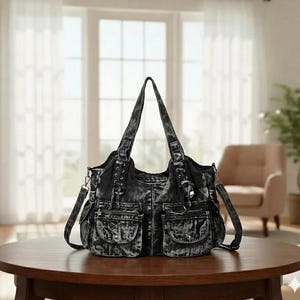May include: A black denim tote bag with multiple pockets and a shoulder strap. The bag has a distressed, faded appearance and is sitting on a round wooden table. The bag has two handles and a zipper closure.