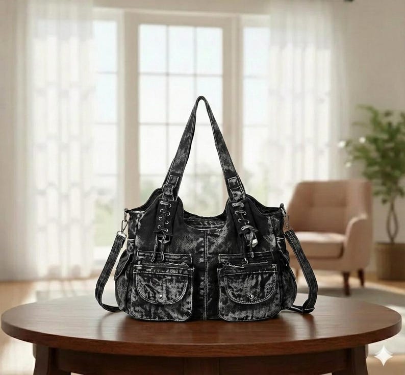 May include: A black denim tote bag with multiple pockets and a shoulder strap. The bag has a distressed, faded appearance and is sitting on a round wooden table. The bag has two handles and a zipper closure.