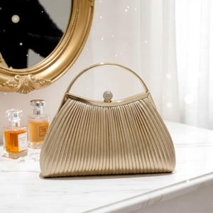 Könnte beinhalten: Eine goldfarbene, plissierte Handtasche mit einem abgerundeten Griff und einem kleinen, dekorativen Schmuckstein oben. Die Tasche hat eine strukturierte Form und steht auf einer weißen Oberfläche. Ein goldgerahmter Spiegel und Parfümflaschen sind im Hintergrund.