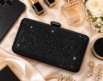 May include: A rectangular black clutch bag covered in small, sparkling crystals with a black gemstone clasp. The bag is positioned on a smooth, cream-colored fabric surface alongside a smartphone, lipstick, perfume bottle, and jewelry.