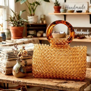 May include: An amber-coloured beaded handbag with a tortoiseshell handle rests on a wooden table. The bag is rectangular with a rounded handle. A small vase of dried flowers and a stack of folded blankets are nearby.