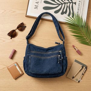May include: A dark blue denim crossbody bag featuring a single shoulder strap, two front zippered pockets with silver-toned hardware, and side pockets. The bag is constructed from durable denim fabric with visible stitching and matching zipper pulls.