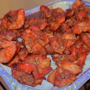 May include: A tray filled with golden-brown fried chicken pieces. The chicken is arranged on a white paper liner inside a silver foil tray. The chicken appears crispy and well-seasoned, with a reddish-brown hue.