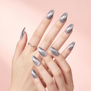 May include: Close-up of hands with long, oval-shaped gray nails. The nails have a shimmering, cat-eye effect, with a stripe of lighter gray running down the center. A silver ring with a wavy design adorns one finger. The background is a soft, pink color.